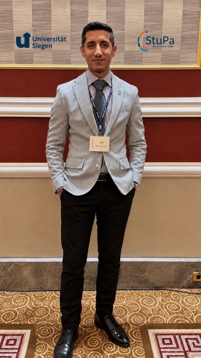 Faizan in a grey blazer with a delegate badge, in front of Universität Siegen and StuPa logos