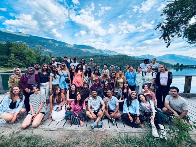 A large group of Erasmus+ participants on a lakeside jetty, with mountains and blue sky behind